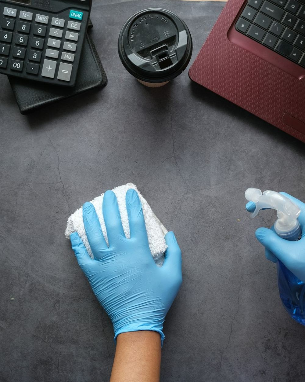 Gloved hands cleaning a desk with spray and cloth